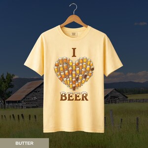 May include: Pale yellow t-shirt featuring a heart made of beer glasses, with the words "I" and "BEER" above and below. The shirt hangs on a wooden hanger, set against a rural landscape with barns and fields.