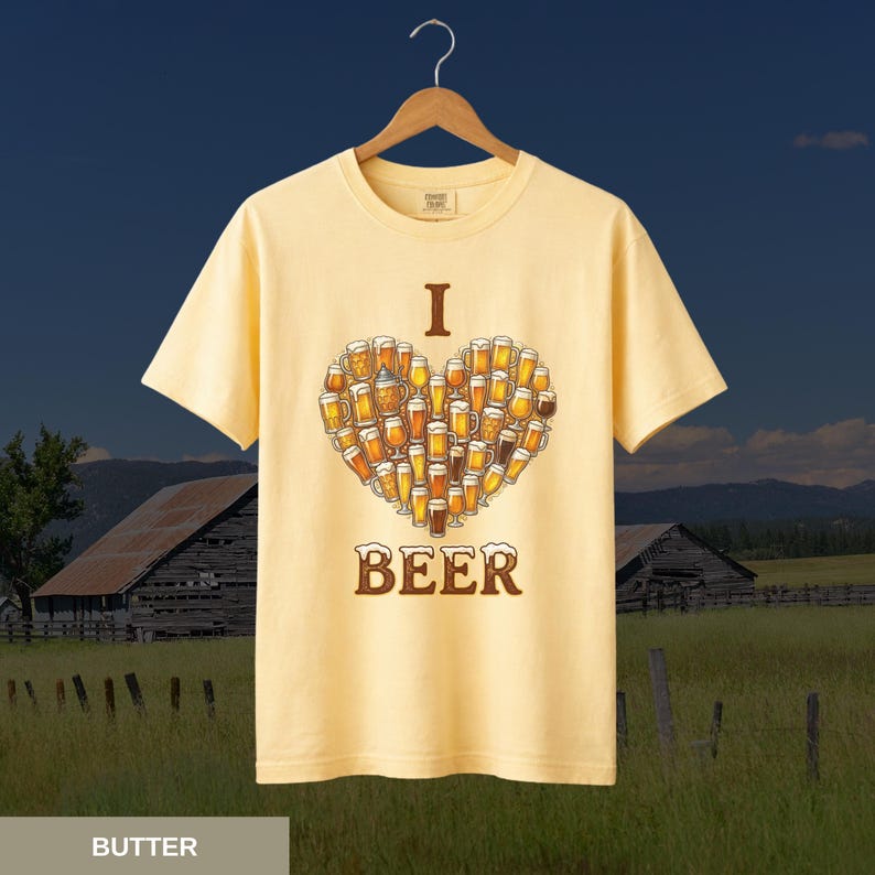 May include: Pale yellow t-shirt featuring a heart made of beer glasses, with the words "I" and "BEER" above and below. The shirt hangs on a wooden hanger, set against a rural landscape with barns and fields.