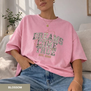 Könnte beinhalten: Hellrosa T-Shirt mit dem Text "DREAMS COME TRUE EST. 2026" in dekorativer Schrift. Das Shirt hat eine lockere Passform. Das Wort "BLOSSOM" befindet sich am unteren Bildrand.