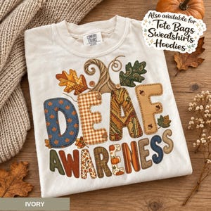 May include: An ivory t-shirt featuring a graphic with the text DEAF AWARENESS. The letters are filled with various autumn-themed patterns including plaid, floral, and denim textures, accented by illustrated pumpkins, acorns, and fall leaves.