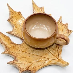 Puede incluir: Un juego de taza y platillo de cerámica. La taza y el platillo son de color marrón cálido, con el platillo en forma de una hoja grande. La taza tiene un asa y se asienta sobre el platillo en forma de hoja. El conjunto está sobre un fondo blanco.