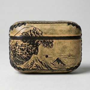 May include: A rectangular, tan-colored box with a lid, featuring a black embossed design of a mountain and a large wave. The box has a dark border and leaf-like accents around the edges, creating a vintage aesthetic.
