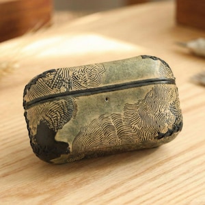 May include: A rectangular, olive-green leather case with a black stitched border. The case features a detailed, wave-like pattern in black. The case is resting on a light-colored wooden surface.