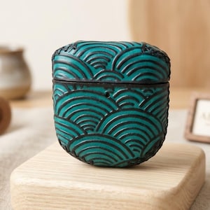 May include: A teal and black protective case for wireless earbuds featuring a repeating embossed wave pattern across the entire surface. The case has a hinged lid design and a smooth, rounded rectangular shape with visible stitching along the edges.