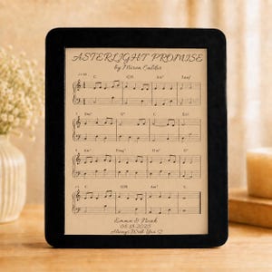 May include: A rectangular black frame containing a beige sheet with printed musical notation titled Asterlight Promise by Miren Caldor. The sheet includes chords, staff lines, and the text Emma & Noah 06.18.2025 Always With You with a heart symbol.