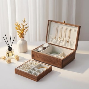 Puede incluir: Una caja de joyería de madera con tapa con bisagras, abierta para revelar compartimentos forrados con tela color crema. La caja contiene collares, relojes y otras joyas. Una bandeja separada más pequeña está al frente, también llena de joyas.