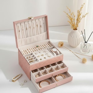 May include: A pink jewelry box with multiple compartments and drawers. The box is open, revealing various jewelry items, including necklaces, rings, and a watch. The box has a gold clasp and is set against a white background.
