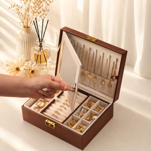 Puede incluir: Un joyero de madera marrón oscuro con tapa abatible que incluye un espejo integrado. El interior está forrado con tela color crema y contiene múltiples ganchos para collares, ranuras acolchadas para anillos y compartimentos para joyas doradas.