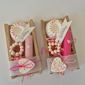 Könnte beinhalten: Zwei kleine Holzkisten gefüllt mit Geburtstagsgeschenken. Jede Kiste enthält einen weiß glasierten Kuchen mit bunten Streuseln, eine rosa Kerze mit einem Herz, Holzperlen und eine herzförmige Dekoration mit Streuseln. Ein Banner lautet "Happy Birthday."