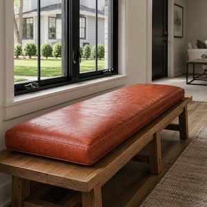 May include: A wooden bench with a rectangular, reddish-orange leather cushion. The bench is made of light-colored wood and is positioned in front of a large window. The cushion appears to be well-padded and provides a comfortable seating area.