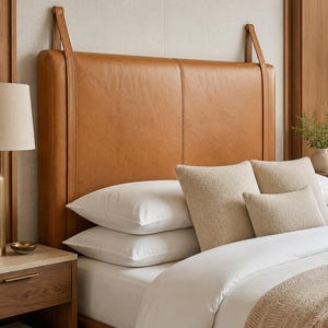 May include: A rectangular tan leather headboard featuring a vertical centre seam and two matching leather straps that extend from the top edge to the wall. The headboard is positioned behind a bed with white pillows and textured beige cushions.
