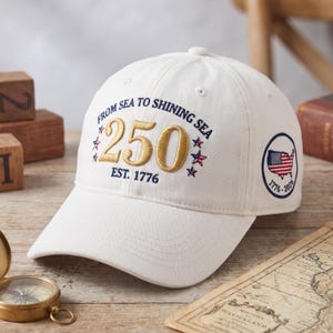 May include: White baseball cap with embroidered text "FROM SEA TO SHINING SEA" and "250 EST. 1776". Features a circular patch with the American flag and the years 1776-2026. A classic, patriotic accessory.