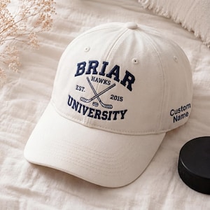 Może przedstawiać: Kremowa czapka bejsbolowa z granatowym napisem "Briar University Hawks", skrzyżowanymi kijami hokejowymi i rokiem 2015. Czapka ma również tekst "Custom Name". Widoczny jest czarny krążek hokejowy.