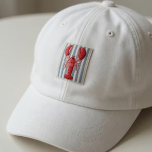 Puede incluir: Gorra de béisbol blanca con un parche bordado de una langosta roja. El parche presenta una langosta roja sobre un fondo a rayas azules y blancas. La gorra tiene una visera curva y un botón en la parte superior.
