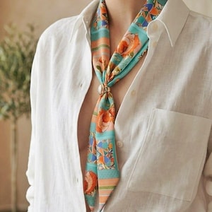 May include: A colorful silk scarf with a floral and geometric pattern is knotted and worn with a white button-down shirt. The scarf features shades of orange, blue, and green. A pearl-like ring secures the knot.