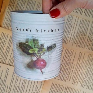 May include: A white metal container with the text "Vera's kitchen" and a radish illustration. The container is held over a background of aged book pages. The radish has a red bulb and green leaves.
