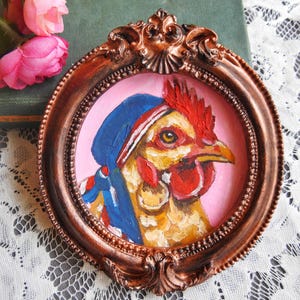 Può includere: Un dipinto a olio raffigurante una gallina con un foulard blu e un orecchino a cerchio dorato, su sfondo rosa chiaro. L'opera è racchiusa in una cornice circolare decorata, color bronzo metallizzato, con dettagli in rilievo.