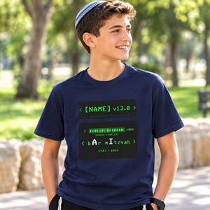 Può includere: Una t-shirt blu navy a maniche corte con una grafica di interfaccia digitale verde. Il design include il testo [NAME] v13.0, una barra di caricamento con la scritta UPDATE COMPLETE 100% e il testo bAr mItzvah 5787 • 2026 in stile codice.
