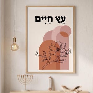 May include: A framed wall art print featuring a black line drawing of a magnolia flower branch layered over terracotta and dusty pink geometric arch shapes on a cream background, with the Hebrew text "עץ חיים" printed in black at the top.