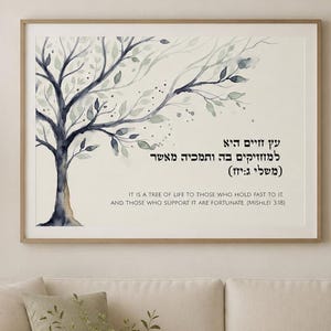 Könnte beinhalten: Ein gerahmter horizontaler Wandkunstdruck mit einem Baum im Aquarellstil mit dunkelblauen und grauen Zweigen und Blättern. Der Text auf Hebräisch und Englisch lautet: It is a tree of life to those who hold fast to it and those who support it are fortunate.