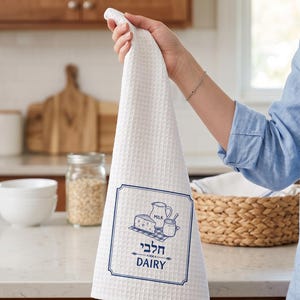 Peut inclure: Un torchon de cuisine blanc à tissage gaufré avec une bordure rectangulaire bleue et une illustration graphique d'un pichet, d'un pot et d'un morceau de fromage. Sous l'illustration, le texte hébreu "חלבי" est imprimé au-dessus du mot "DAIRY".