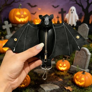 May include: A black bat-shaped coin purse made of textured material with white stitching along the wings. The centre features a vertical silver-toned metal zip and a matching silver-toned lobster clasp attached to the bottom for hanging.