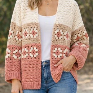 May include: A cream, tan, and dusty rose crochet cardigan. The open-front sweater features a granny square design with a solid color band at the bottom of the sleeves and the bottom of the cardigan. The sleeves are three-quarter length.