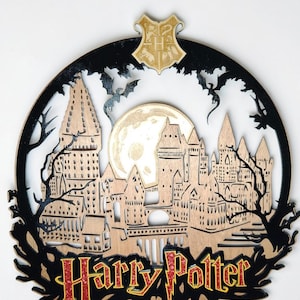 Może przedstawiać: Drewniana dekoracja ścienna z motywem Harry'ego Pottera przedstawiająca szczegółową sylwetkę zamku Hogwart, pełnię księżyca i nietoperze. Napis "Harry Potter" jest w czerwonym i złotym brokacie. Złoty herb na górze.