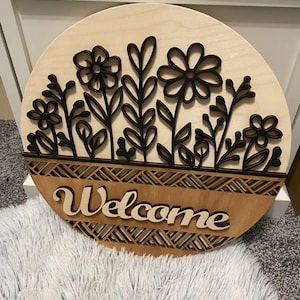 Puede incluir: Un letrero redondo de madera con un diseño floral y la palabra "Welcome". El letrero presenta flores y hojas cortadas con láser en negro sobre un fondo de madera clara. La palabra "Welcome" está en una fuente cursiva.