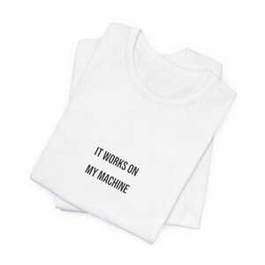 Peut inclure: Deux t-shirts blancs pliés et empilés. Le t-shirt du dessus porte l'inscription "IT WORKS ON MY MACHINE" en noir. Les chemises sont en matière souple, probablement en coton ou en mélange de coton.