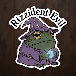 May include: A sticker featuring a green frog wearing a purple wizard hat with yellow stars and moons, and a purple robe tied with a rope belt. The frog holds a glowing blue orb. The text Rizzident Evil is printed in black above the frog.