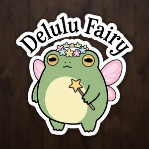 May include: A die-cut sticker featuring a green frog with a cream-coloured belly, wearing a pastel flower crown, and sporting pink fairy wings. The frog holds a yellow star-tipped wand. Above the frog, the text reads Delulu Fairy in black.