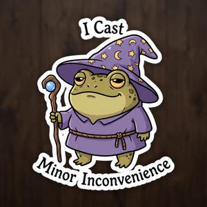 May include: A sticker featuring a green toad wearing a purple wizard hat with yellow stars and moons, and a purple robe with a rope belt. The toad holds a brown wooden staff with a blue orb. The text reads I Cast Minor Inconvenience.