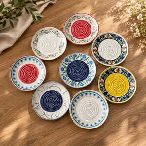 May include: Eight round ceramic grater plates featuring textured centers for grating and decorative floral borders in various colors including red, blue, yellow, and white. Each plate has a distinct pattern and color scheme on the rim.