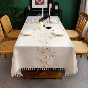 Può includere: Una tovaglia rettangolare color crema con un motivo floreale ricamato in oro e azzurro. La tovaglia è appoggiata su un tavolo di legno con sedie abbinate. I bordi della tovaglia sono rifiniti con nappe. La tavola è apparecchiata con piatti, bicchieri e candele.