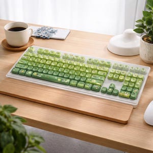 Peut inclure: Un clavier mécanique blanc avec des touches vertes. Les touches présentent un dégradé de nuances de vert et de petites illustrations. Le clavier est posé sur un bureau en bois avec un repose-poignet en bois.