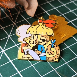 Peut inclure: Épinglette en émail colorée représentant un personnage de dessin animé avec un chapeau de paille, une chemise bleue et un serpent. La broche a une bordure et un dos dorés. Le personnage tient une tasse.