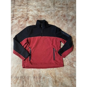 May include: A black and red fleece pullover with a high collar and a quarter-zip. It features two zipped side pockets and a small sleeve pocket. The sleeves and upper chest are black, the lower body is red. A warm garment.