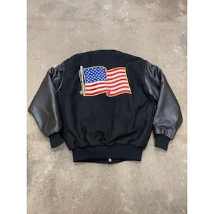May include: Black varsity jacket with leather sleeves. The back features an embroidered American flag in red, white, and blue. The jacket has a ribbed collar, cuffs, and hem.