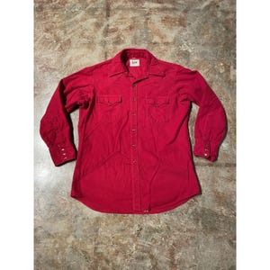 May include: A red long-sleeve shirt with a button-down front, two chest pockets, and a pointed collar. The shirt has buttoned cuffs and a small Lee label on the collar. The shirt is made of a soft, possibly cotton, material.
