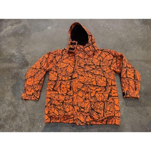 Puede incluir: Chaqueta de caza naranja con un patrón de ramas de árboles negras. La chaqueta tiene capucha, cierre de cremallera y bolsillos. La chaqueta está hecha de un material duradero.