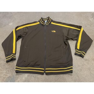May include: A brown zip-up track jacket with yellow stripes on the sleeves and around the collar and waist. The North Face logo is embroidered on the chest. The jacket has two front pockets.
