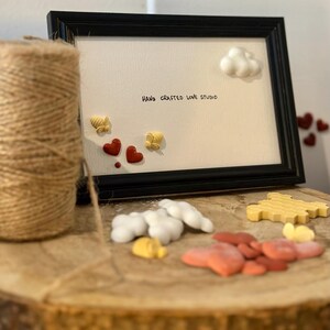 May include: A black framed art piece with the text "HAND CRAFTED LOVE STUDIO" and decorative elements including red hearts, white clouds, and yellow honeycomb shapes. A roll of twine sits to the left.