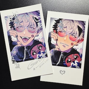 May include: Two polaroid-style prints featuring anime-style illustrations of a character with white and black hair, wearing glasses and a black jacket. The character has a playful expression in one print and a more serious expression in the other. Each print has a unique design element.