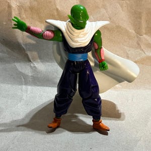 May include: A green action figure of the character Piccolo from Dragon Ball Z. The figure is wearing a white cape, blue pants, and orange boots. The figure has a stern expression and is posed with one arm outstretched.