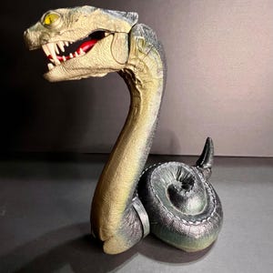 May include: A toy snake with a detailed design. The snake has a tan and green body, with a black tail. The head is open, revealing white teeth and a red mouth. The eyes are yellow.