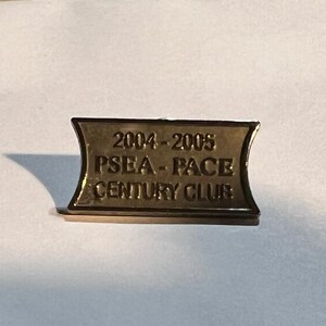 May include: A rectangular gold-toned metal lapel pin with concave sides. The surface features embossed text that reads 2004-2005, PSEA-PACE, and CENTURY CLUB. The pin has a polished metallic finish and a distinct border framing the lettering.