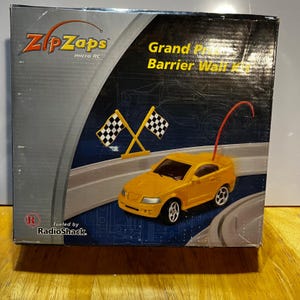 May include: A ZipZaps micro RC Grand Prix Barrier Wall Kit box featuring a yellow toy car with a red antenna, two checkered flags, and the RadioShack logo. The packaging displays a blue and black background with a white track graphic.