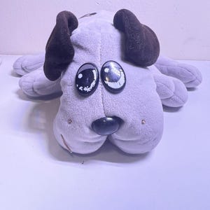 May include: A plush dog toy in a light gray color with dark brown ears. The dog has large, oval-shaped eyes and a black nose. The toy is lying down, with its paws outstretched.