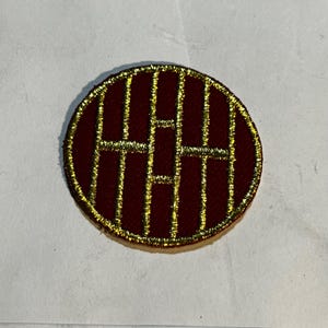May include: A circular embroidered patch featuring a deep maroon background with a metallic gold thread design. The pattern consists of four vertical lines intersected by two horizontal bars, all bordered by a thick gold stitched edge.